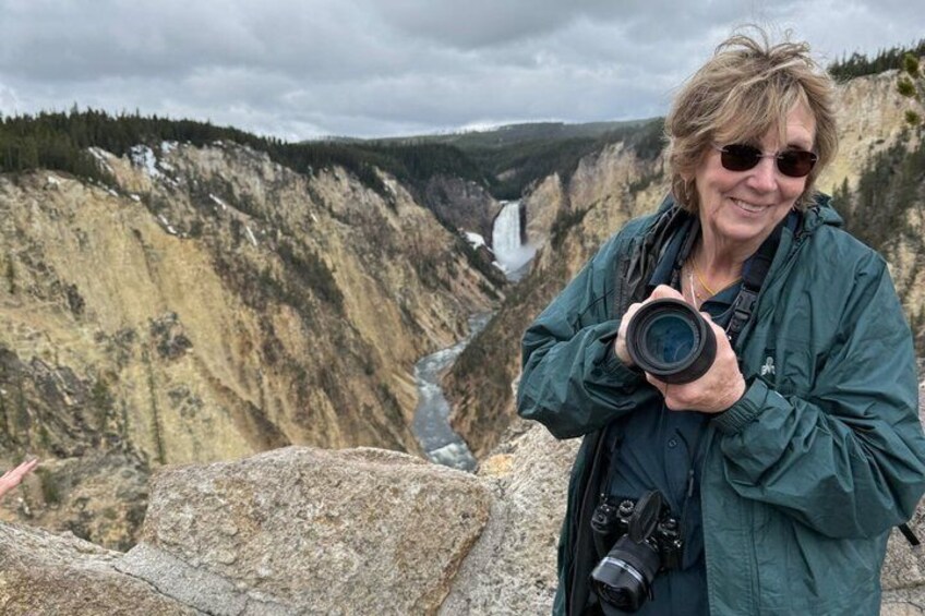 A person holding a camera, standing in front of a canyon with a river and waterfall. 4