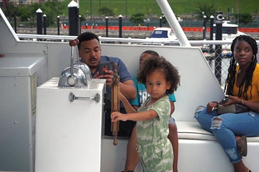 A boat with a steering wheel, a child holding a wooden stick, and adults sitting on the deck. 4
