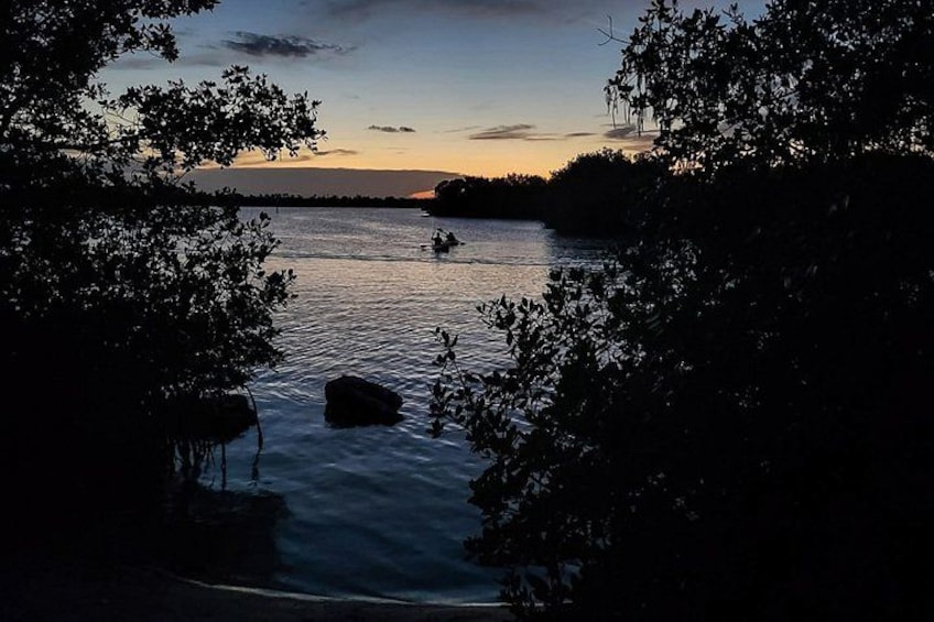 A calm lake at dusk with trees framing the view, a small boat in the water, and a distant shoreline. 1