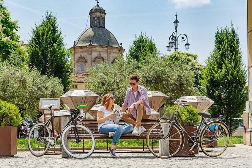 A couple sitting on a bench with bicycles parked nearby, a historic building in the background, and trees around. 5