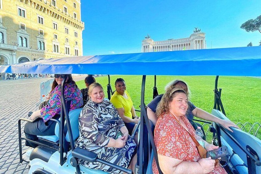 Eine Gruppe von Personen in einem Golfwagen in einem Park, im Hintergrund ein großes Gebäude. 4
