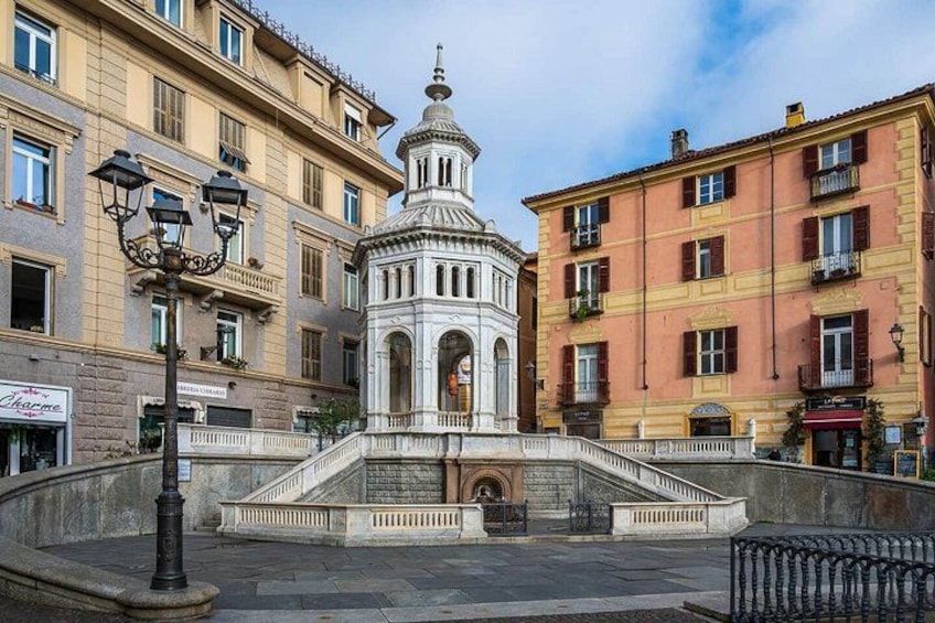A historic fountain with a central structure, surrounded by multi-story buildings with balconies and storefronts. 3