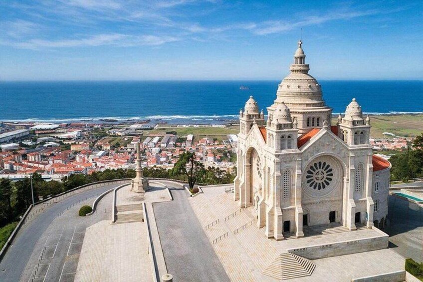 Una gran catedral blanca con una cúpula y un ventanal circular, rodeada de un área pavimentada y vistas al océano y al paisaje urbano. 1