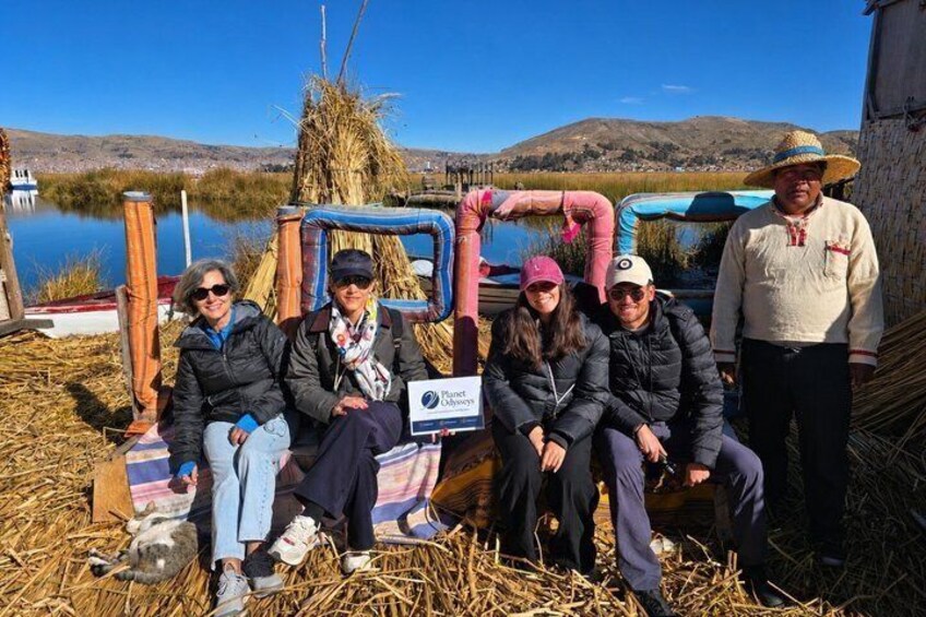 A group of people sitting and standing near a rustic structure by a lake with mountains in the background. 5