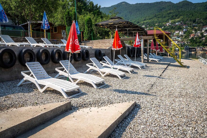 A beach with white lounge chairs and red umbrellas, a wooden structure, and a hillside in the background. 5