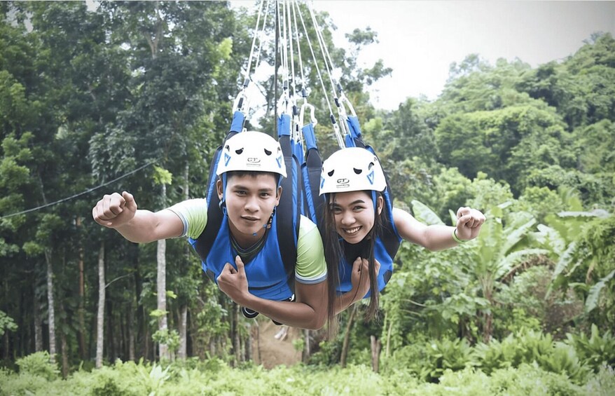 Two people bungee jumping with helmets and harnesses, surrounded by dense forest. 3