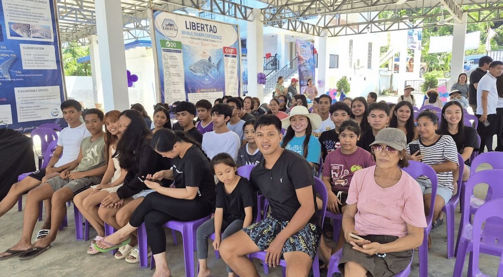 A group of people seated on purple chairs under a covered area with informational posters about a whale shark experience. 2