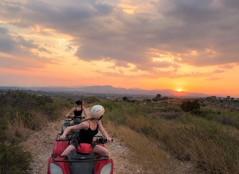Two people on a quad bike on a dirt path with a sunset in the background. 1