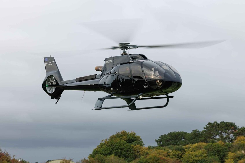 A black helicopter with 'PH-ZUT' on its tail, flying over a landscape with trees and a cloudy sky. 1