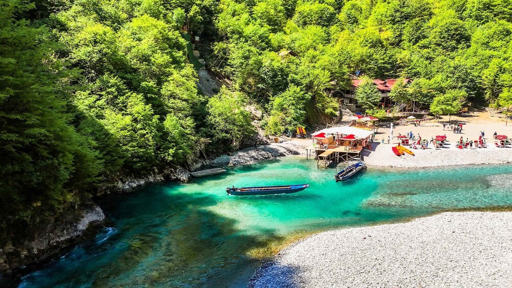A turquoise river with boats, a pebbled beach, and a forested area with buildings. 1