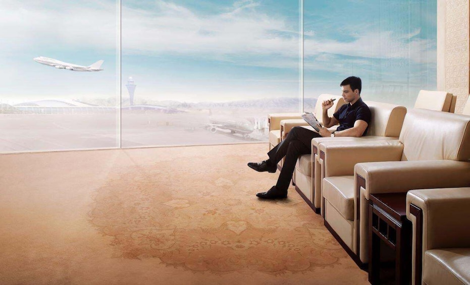 A man sitting on a leather couch in a spacious room with a large window overlooking an airport. 2