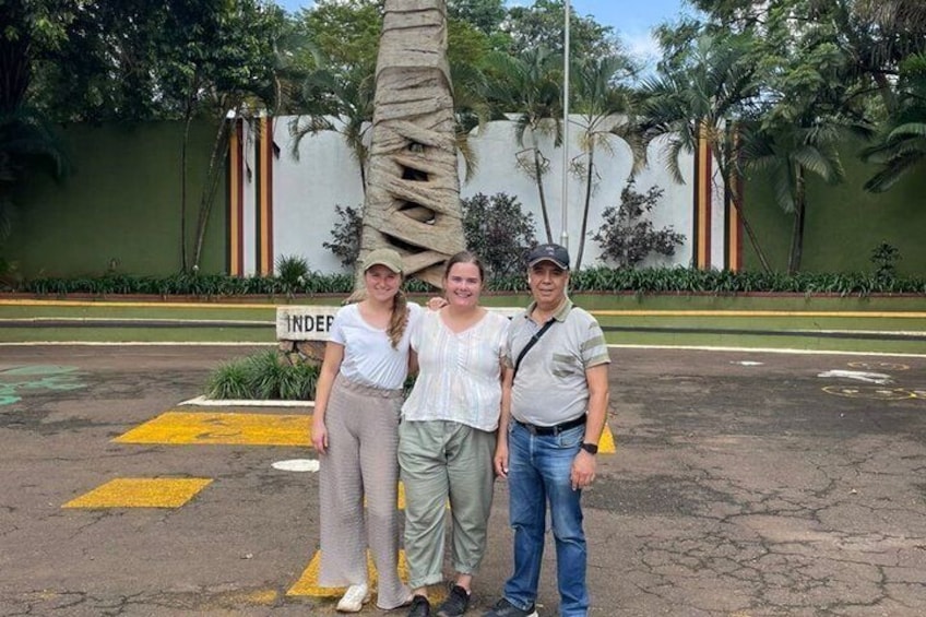 Three people standing in a parking lot with a palm tree and a sign that reads 'INDEF' in the background. 3