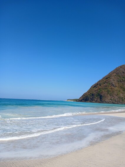 A beach with clear blue water, a sandy shore, and a hill in the background. 4