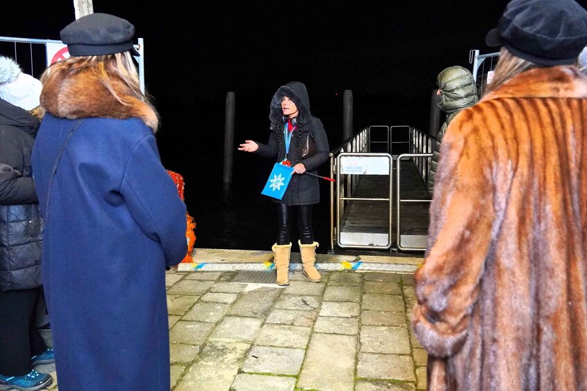 A person in a black coat with a fur-lined hood and a blue bag, standing on a paved area with a metal barrier in the background. 4