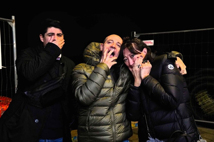 A group of people in winter clothing, some with their hands over their mouths, standing in a dark outdoor setting. 1