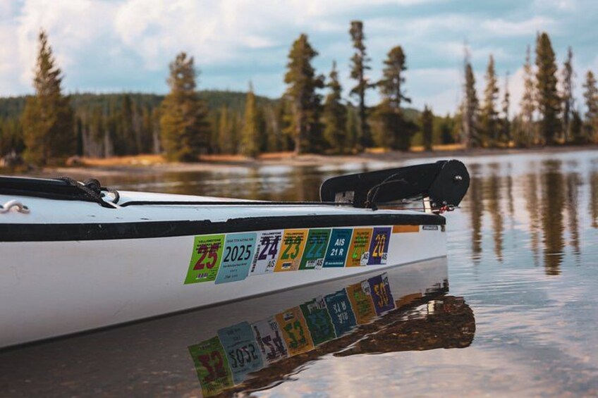 A canoe with stickers on its side, floating on calm water with a forested shoreline in the background. 1