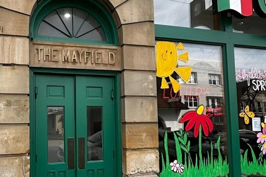A building with a green door labeled 'THE MAYFIELD' and a window decorated with a sun and flowers. 4