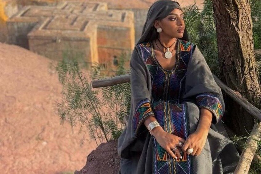 A person sitting on a wooden bench in a desert-like environment with sparse vegetation and a stone structure in the background. 4