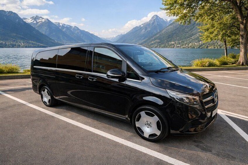 A black minivan parked on a road with a lake and mountains in the background. 3