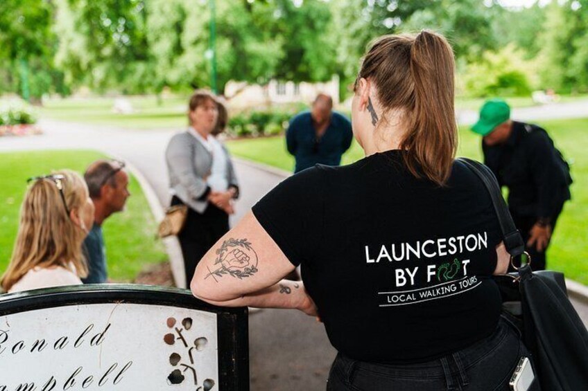 A person wearing a black t-shirt with 'LAUNCESTON BY FOOT' printed on it, standing outdoors with a group of people in the background. 2