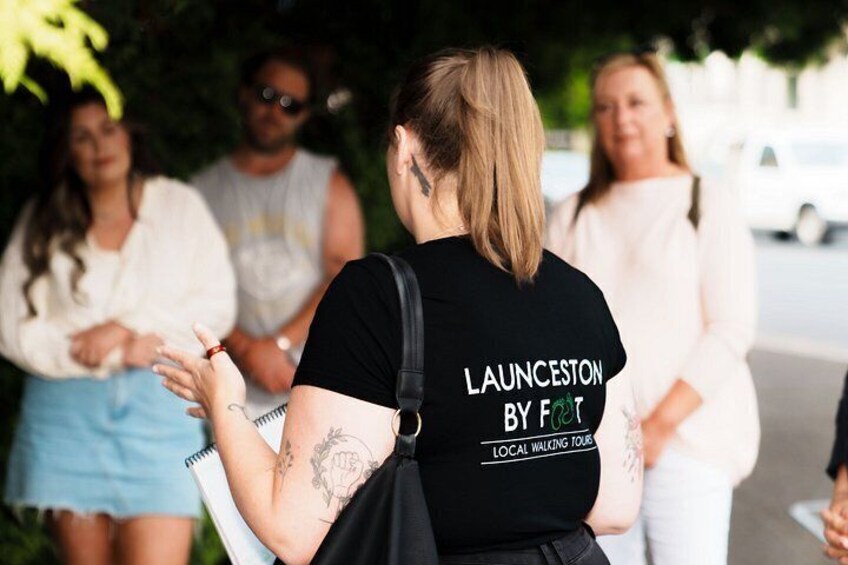 A person wearing a black t-shirt with 'LAUNCESTON BY FIT' printed on the back, holding a notebook. 5