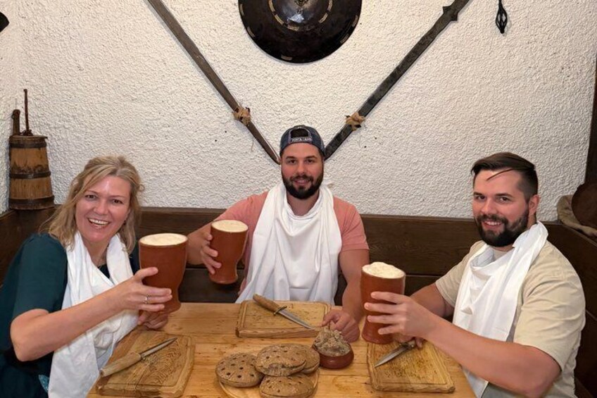 Three people sitting at a wooden table with bread and a knife, in a rustic setting with a barrel and a wall-mounted object. 1