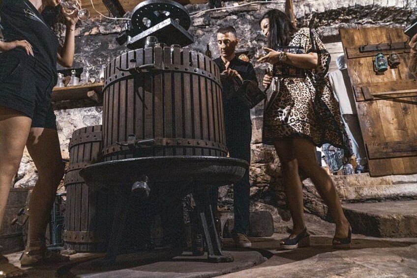 A group of people around a large, cylindrical, industrial-looking structure, possibly a distillation apparatus, in an outdoor setting with stone walls and rustic wooden doors. 3