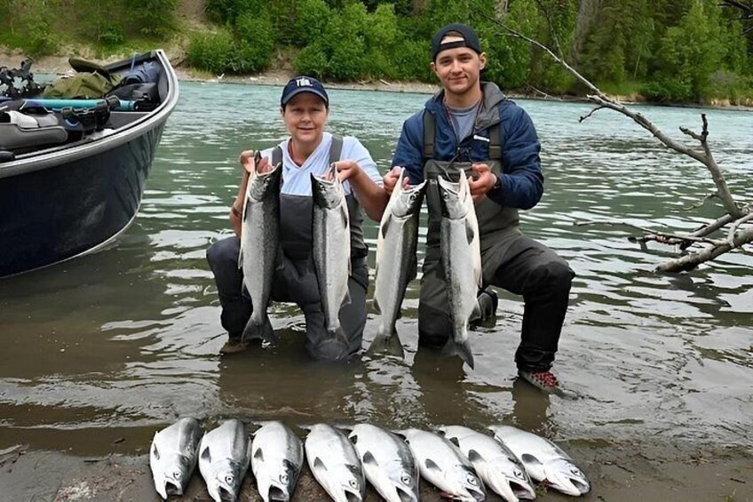 Two people holding fish in a river with a boat and several fish on the ground. 4