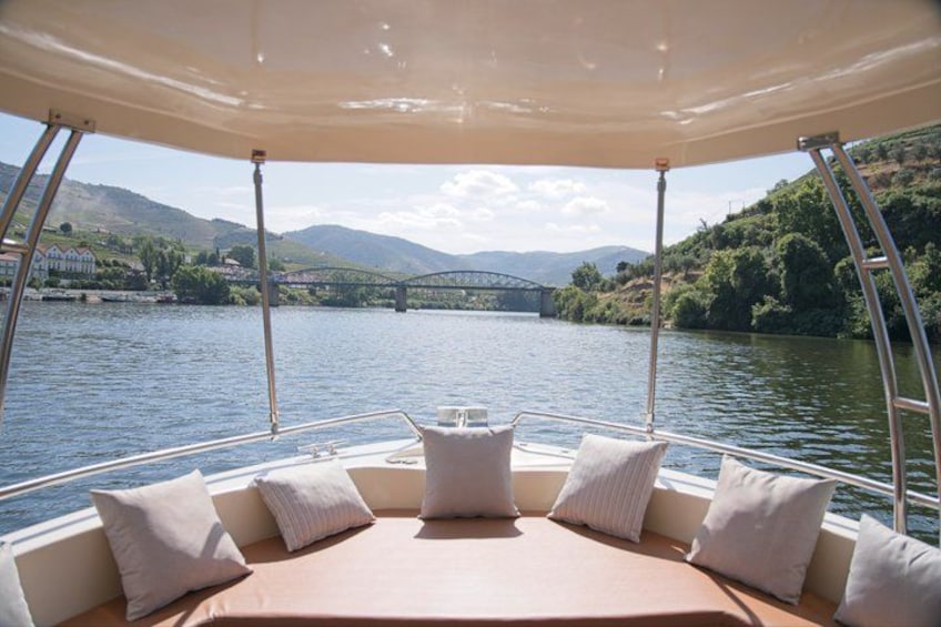 A boat with a covered seating area, a table, and a view of a river with a bridge and hills in the background. 4
