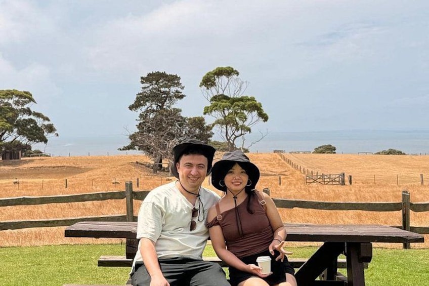 A couple sitting at a picnic table in a grassy field with trees and a fence in the background. 3