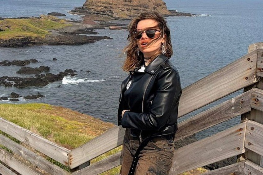 A person standing on a wooden boardwalk by the sea, with rocky cliffs and waves in the background. 5