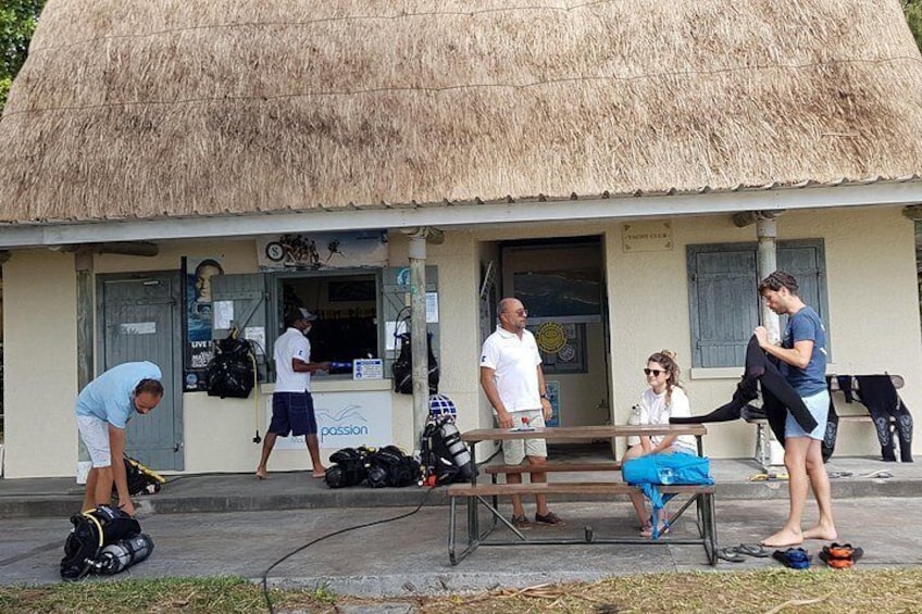 A thatched-roof building with people around it, some sitting and others standing, and bags and equipment on the ground. 4