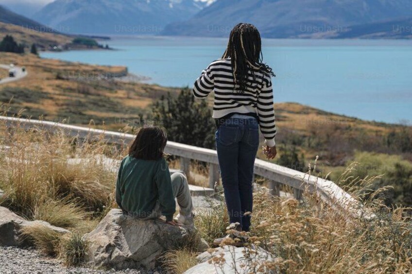 Two people sitting and standing on a rocky outcrop overlooking a lake and mountains. 5