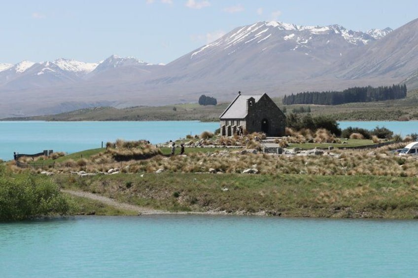 A small stone building near a turquoise lake with mountains in the background. 1