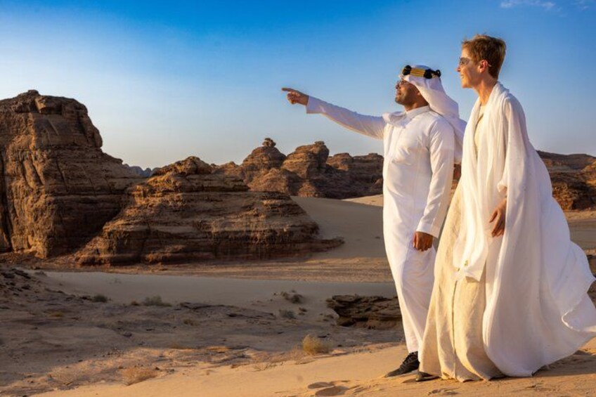 Two people in white robes standing on a desert with rocky formations in the background. 1