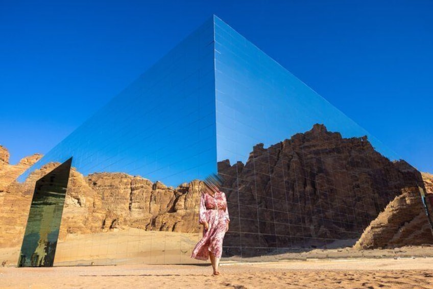 A person in a pink dress walking in a desert with a large glass structure and a mountain in the background. 1
