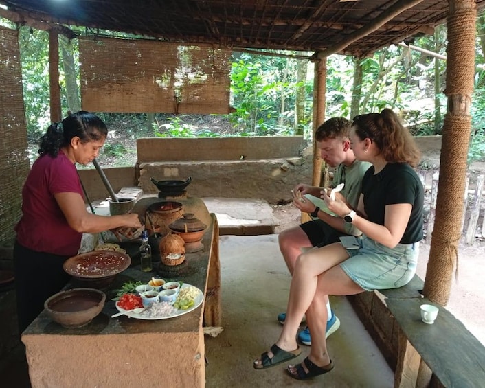 A rustic outdoor dining area with a thatched roof, a wooden table, and a variety of food items and utensils. 5