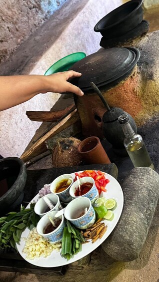 A traditional cooking setup with a mortar and pestle, a plate with various ingredients, and a hand reaching for a lid. 1
