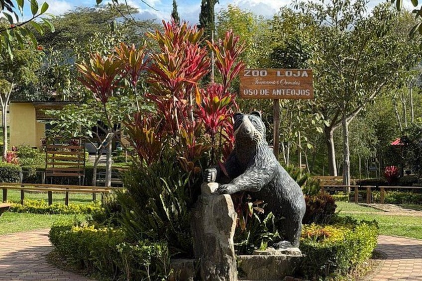 A zoo entrance with a bear statue, lush greenery, and a signboard. 1