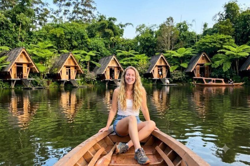 A person in a wooden boat on a calm river, surrounded by forested areas and several wooden houses on stilts. 2