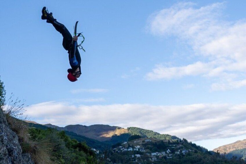 A person in a headstand position with a red helmet, suspended in mid-air against a backdrop of a mountainous landscape with a clear sky. 1