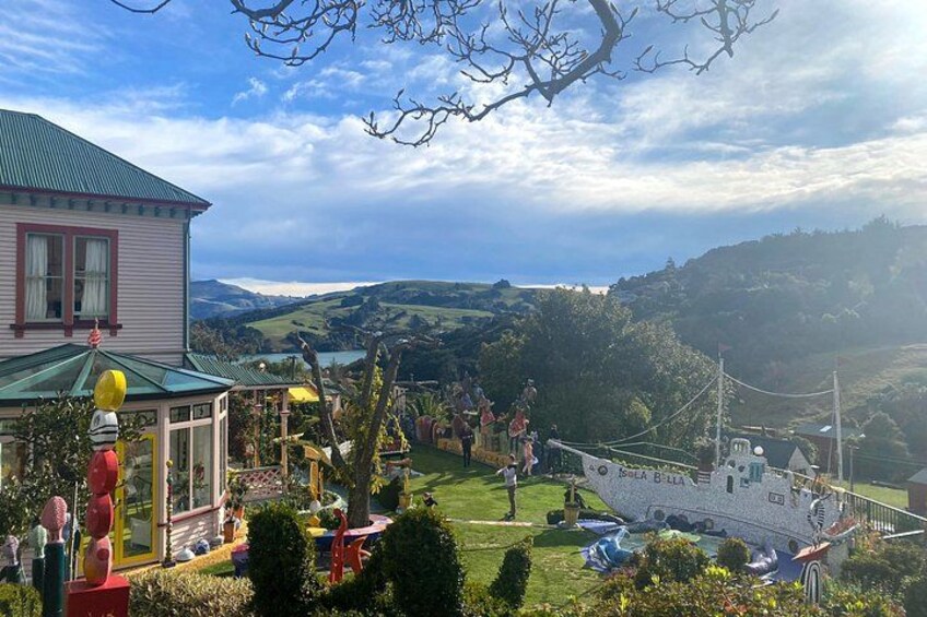 A house with a green roof, a garden with various sculptures, and a boat named 'Korea Balla' in the background. 2