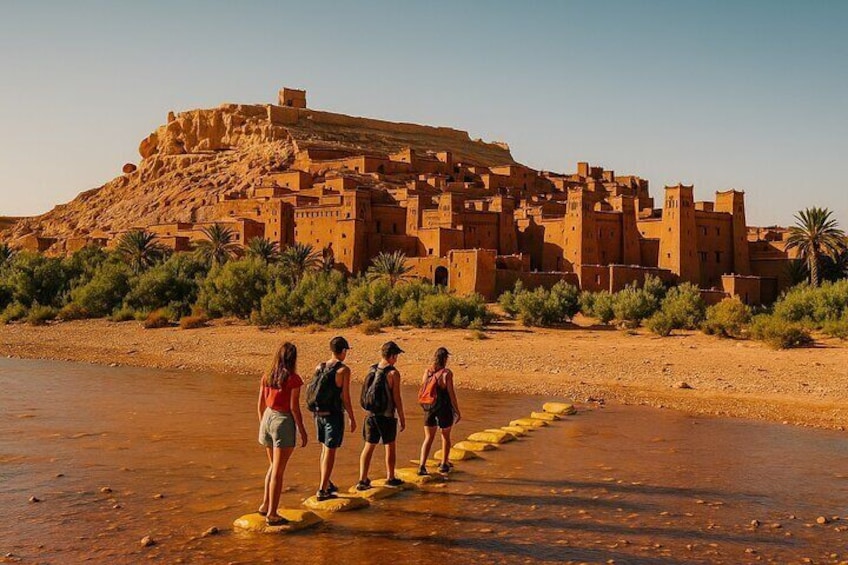 A group of people walking on stepping stones across a river towards a large, ancient mud-brick building. 2
