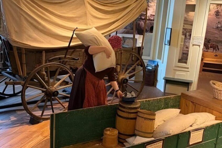 A historical reenactment scene with a person in period costume, wooden wheels, barrels, and a green display case. 2
