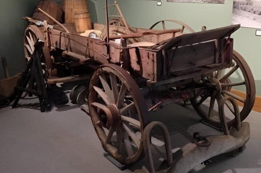 An old wooden carriage with large wheels and a bench seat, displayed in a museum. 5