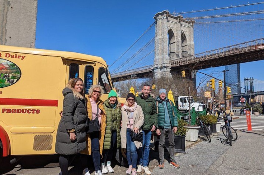 A group of people standing in front of a yellow bus with 'Old Fulton Dairy' written on it, with a bridge in the background. 2