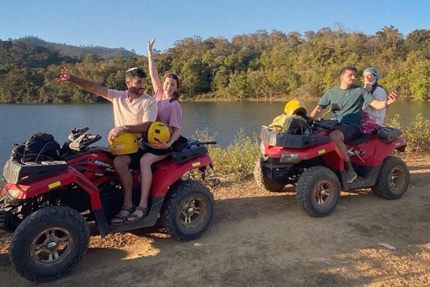 Dos personas en un cuatrimoto rojo, una usando un casco amarillo, cerca de un lago con árboles al fondo. 1