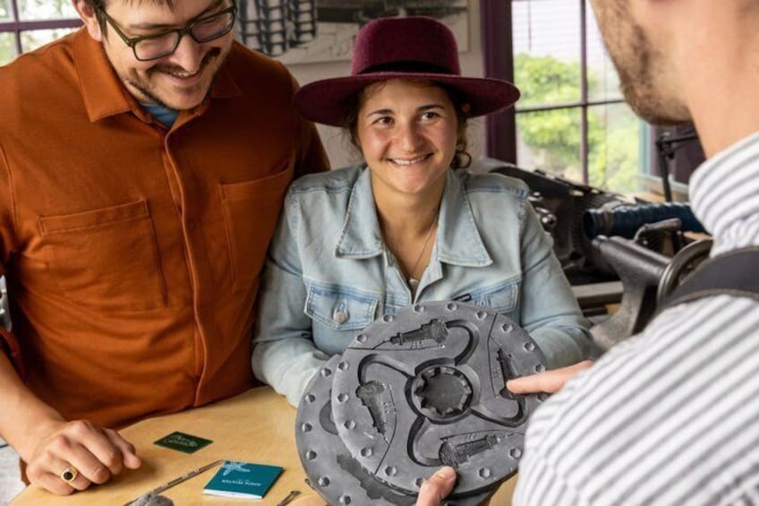 Three people examining a mechanical part with a circular design, a toolbox, and a window in the background. 1