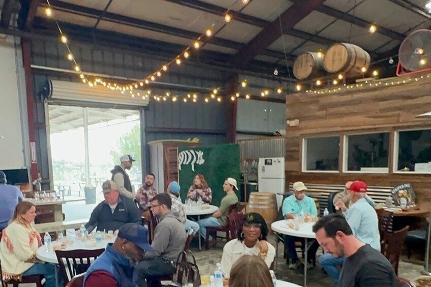 A group of people seated at tables in a rustic indoor setting with string lights and wooden barrels. 3