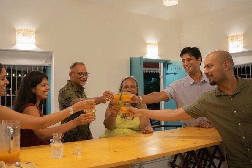 A group of people toasting with drinks at a wooden table in a room with white walls and a blue door. 3